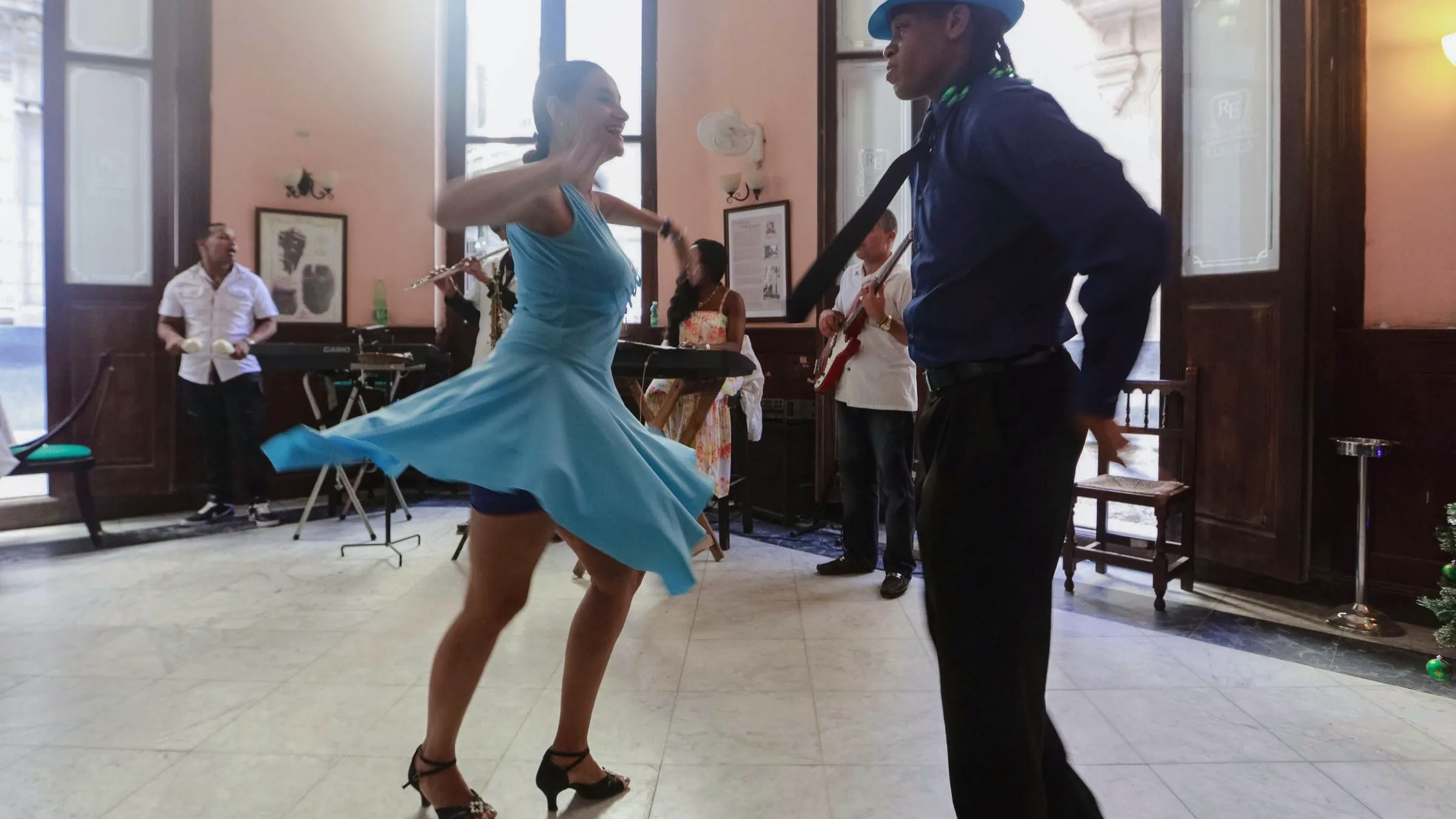 A couple dancing salsa across a tiled floor while musicians play by the windows