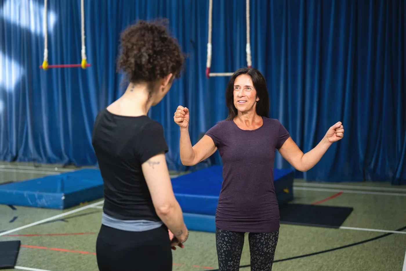Two adult coaches talking on a gym floor beside landing mats, trapezes hanging behind