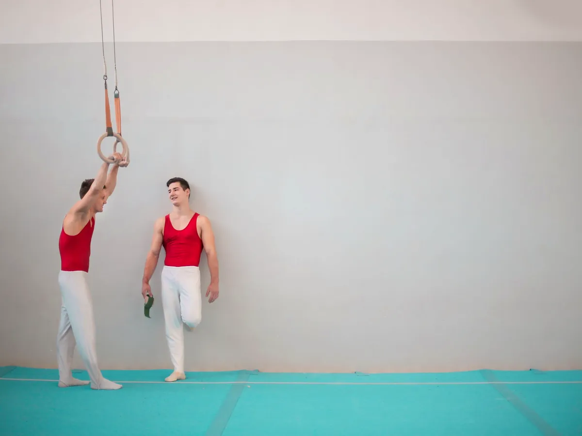 Two adult gymnasts standing by a pair of rings in a bright gym