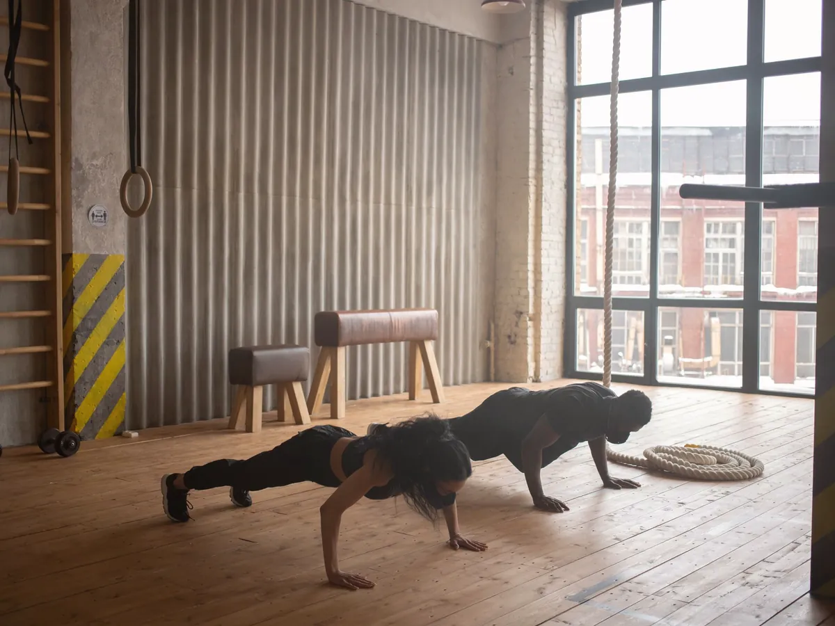 Two adults doing push-ups on a wooden floor in a loft gym