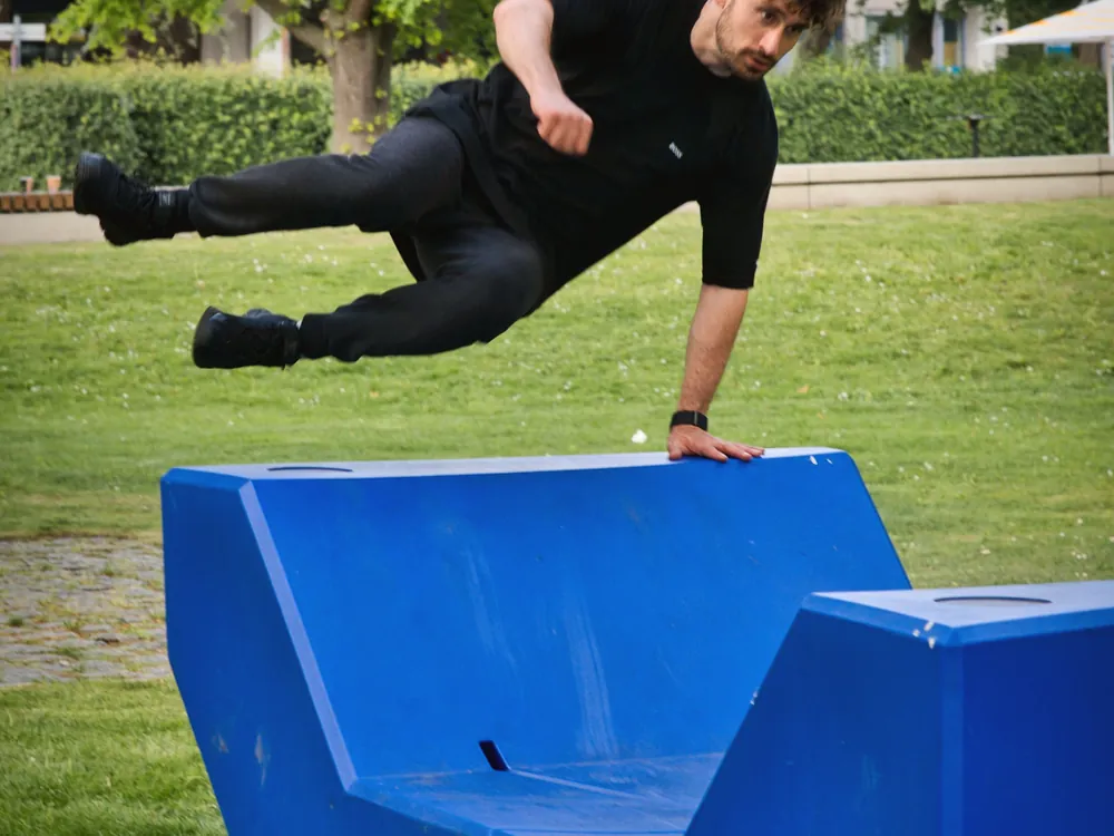 An adult in a black t-shirt vaulting a blue concrete block in a park